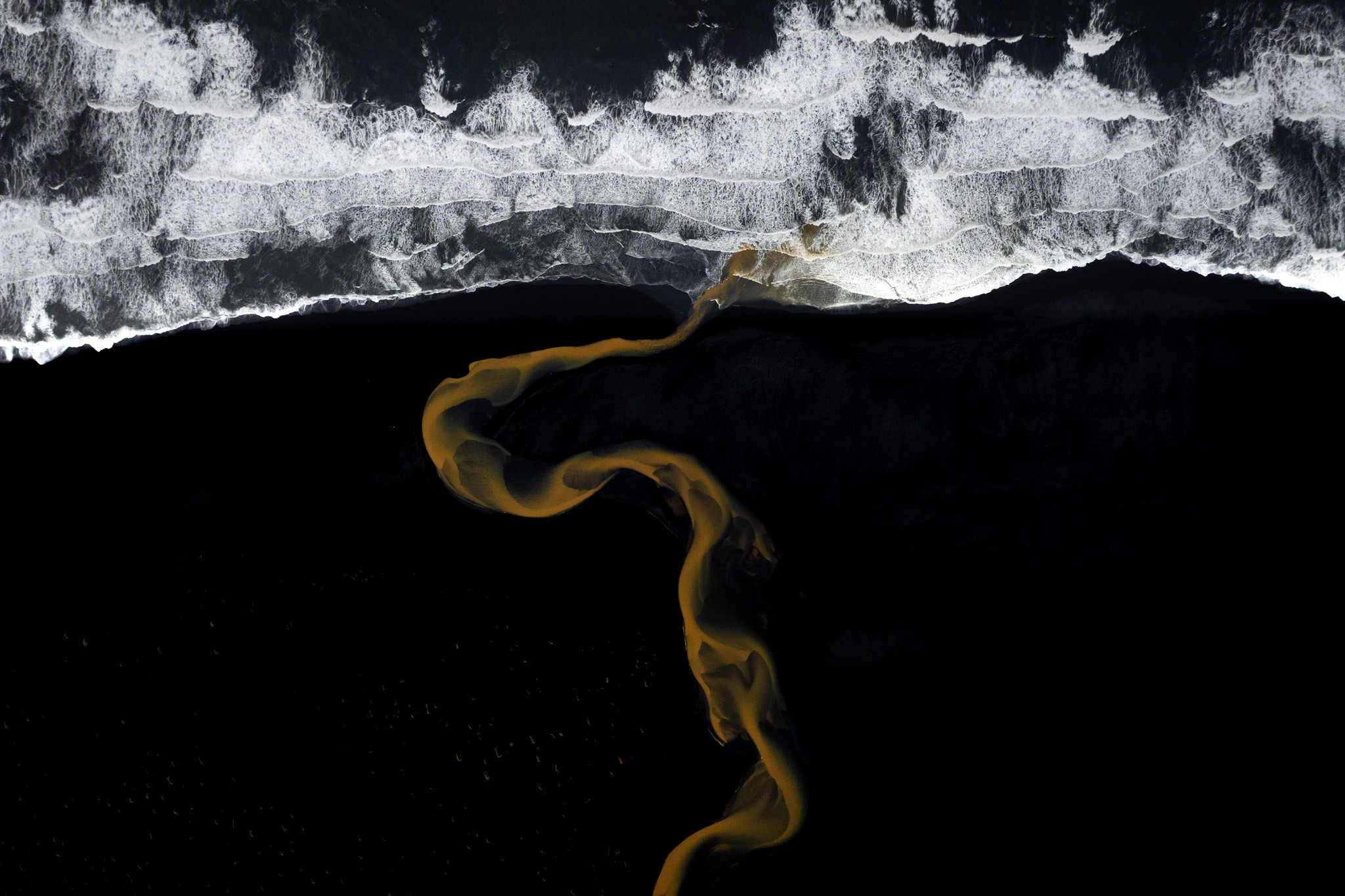 Flowing Forms. This image captures the raw beauty of Iceland's coast from high above. A river, colored by the earth it carries, cuts a path through the wide beach. The sand here is pitch black, created by volcanoes long ago. Against this dark ground, the water looks just like flowing liquid gold. You can see it twisting and turning until it finally reaches the sea. There, the golden water meets the rough, white waves of the ocean. The photo does not rely on many different colors to tell a story. Instead, it uses just three powerful tones: gold, black, and white. This simple mix makes the contrast very intense and draws your eye directly to the shapes and the way the water moves across the land.